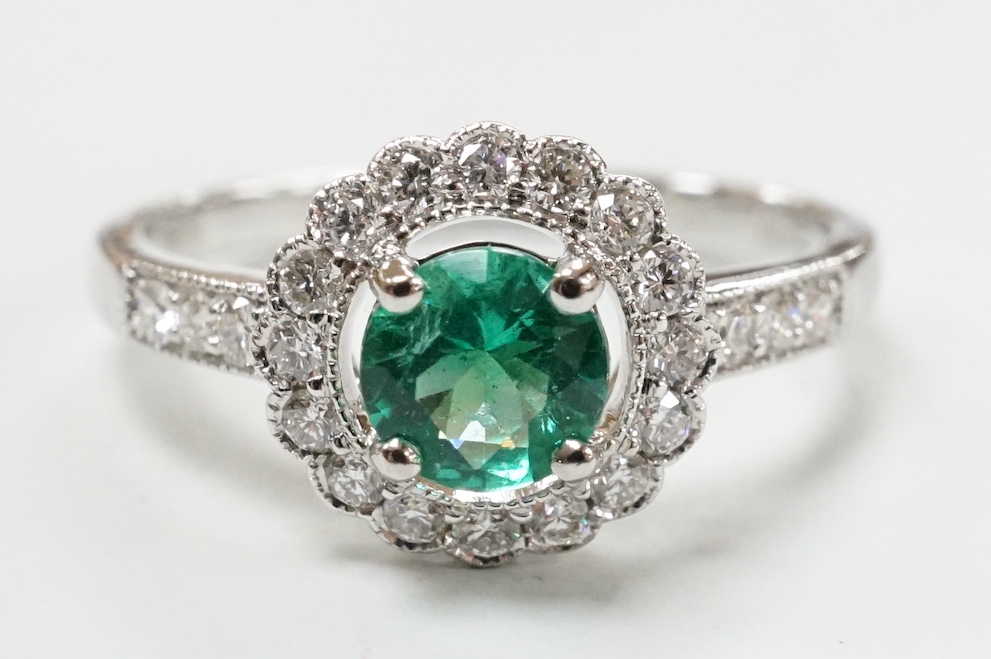 A modern 18ct white gold, emerald and diamond set circular cluster ring, with diamond set shoulders, size M, gross weight 2.7 grams.